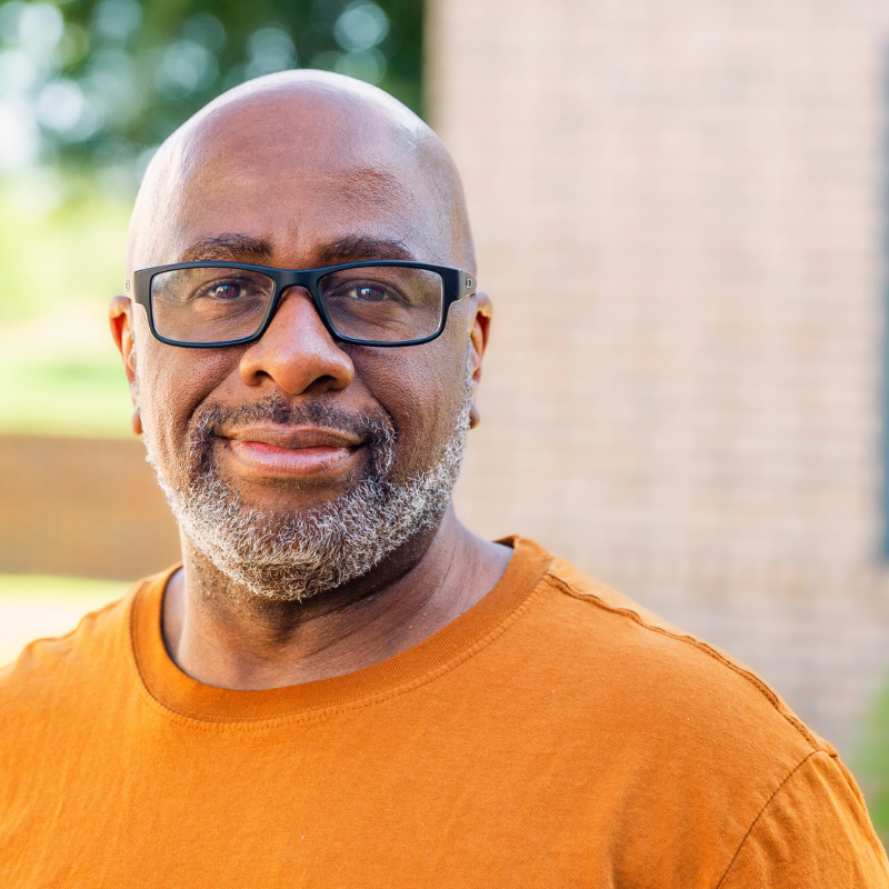 Smiling Black man with glasses in an orange t-shirt outdoors, looking at the camera.