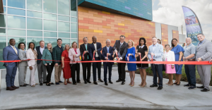 Dignitaries and community members line up outside a colorful modern building for a ribbon‑cutting ceremony, each with scissors to cut a red ribbon.