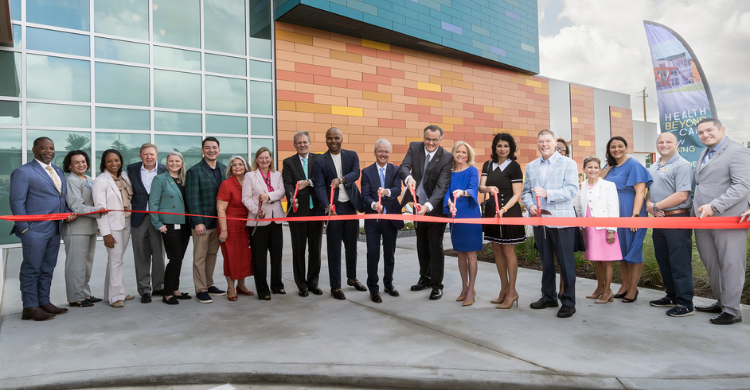 Dignitaries and community members line up outside a colorful modern building for a ribbon‑cutting ceremony, each with scissors to cut a red ribbon.