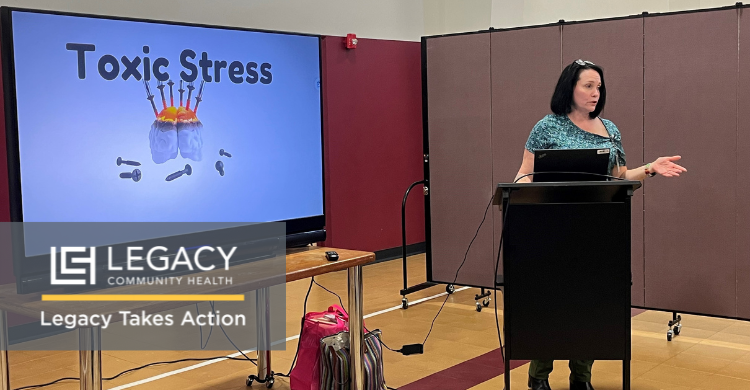 Presenter at a lectern delivering a talk on toxic stress; slide reads 'Toxic Stress' with a brain illustration beside a few scattered items; Legacy branding visible.