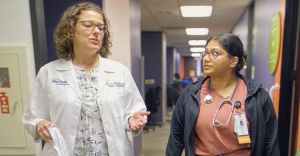 Two female healthcare professionals walk and talk in a hospital corridor, one in a white lab coat and the other wearing a stethoscope and badge.