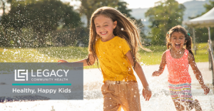 Two smiling kids running through water jets in a sunny park, with a Legacy Community Health logo and the slogan 'Healthy, Happy Kids' overlaid on the image.