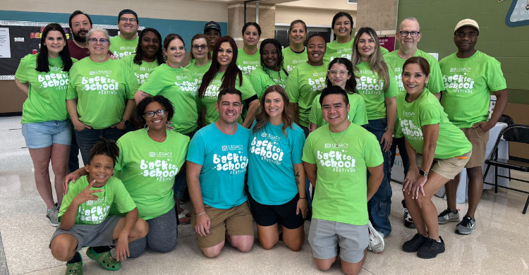 Group of diverse volunteers in bright green 'Back to School Festival' shirts pose together in a school hallway, with a few people wearing blue shirts in the center.