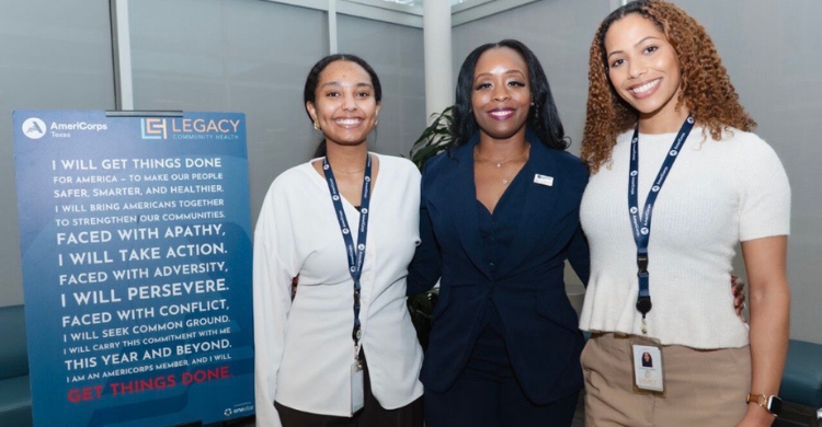 Three women stand together at a conference, smiling, each wearing a lanyard; a blue AmeriCorps poster is visible on the left.