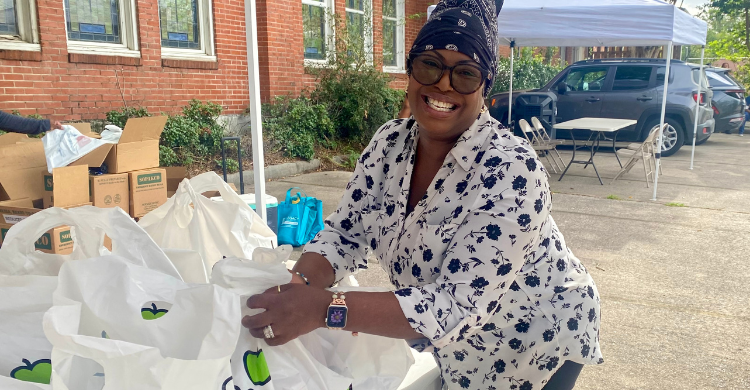 Smiling woman wearing a headscarf and floral shirt packs white grocery bags at an outdoor charity event.