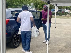 Two men loading bags into a dark SUV under a white canopy in a parking lot, one at the open rear door and the other facing him.