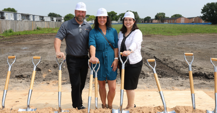Three people in hard hats stand with shovels at a groundbreaking ceremony on a construction site, smiling for the photo.