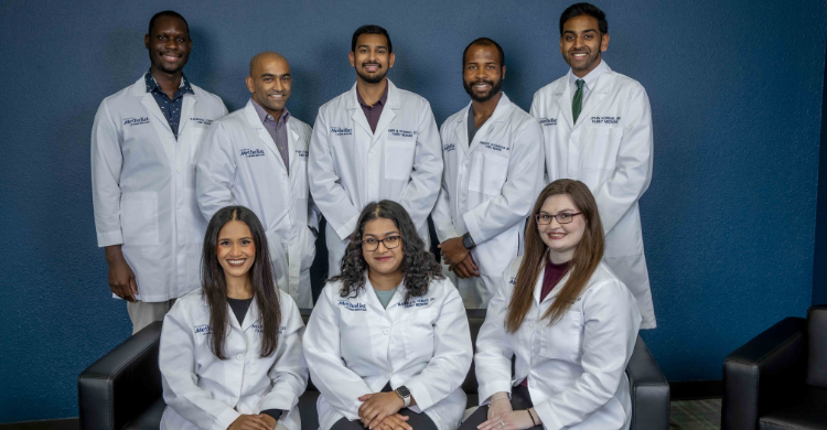 Seven medical students in white coats pose for a team photo in a blue room with seating visible.