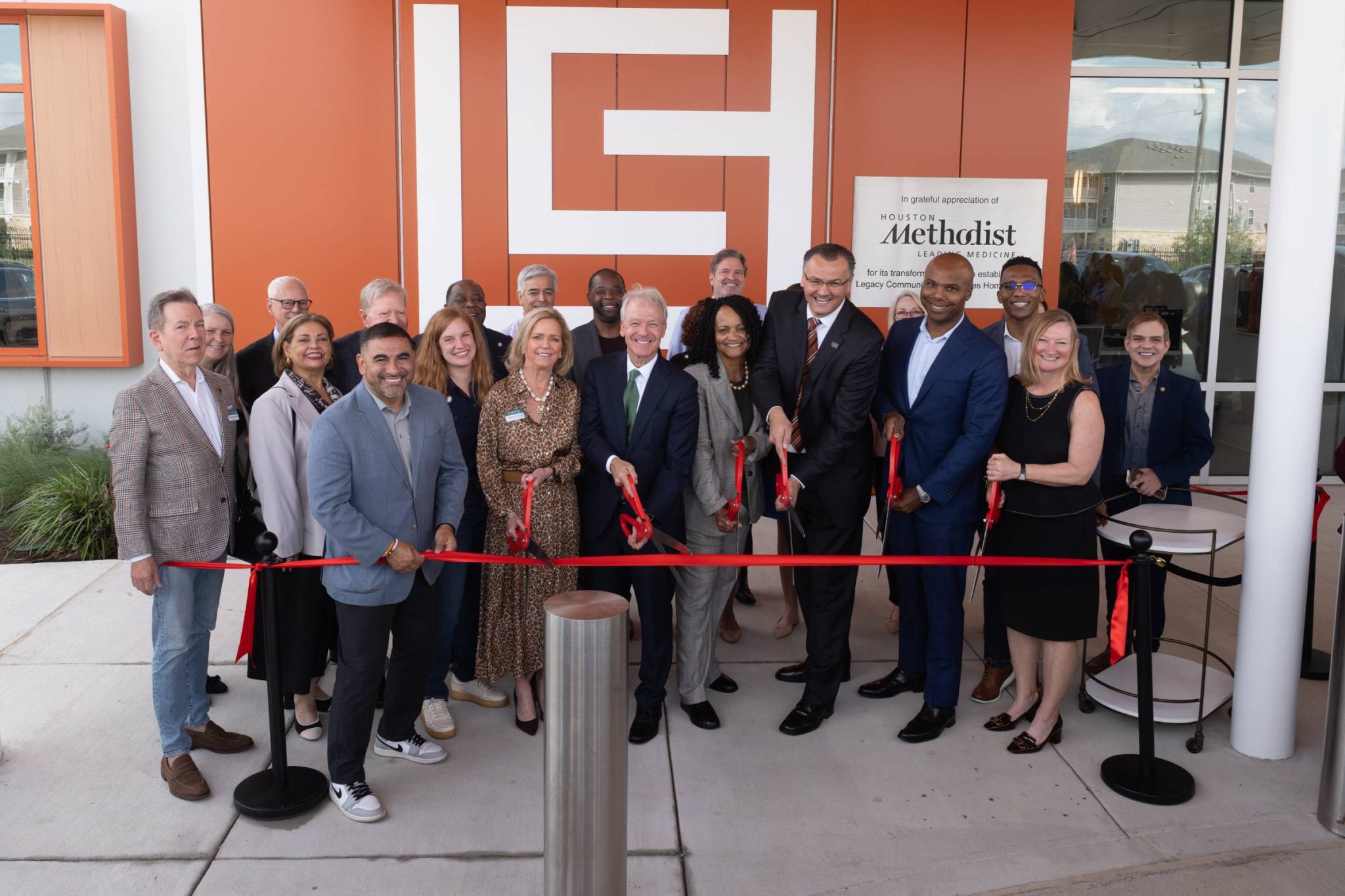 Group of community leaders, elected officials, healthcare leaders and partners participate in a ribbon-cutting ceremony outside the Legacy Community Health Acres Homes Clinic in Houston. Participants stand behind a red ribbon in front of the clinic entrance and Houston Methodist recognition signage.