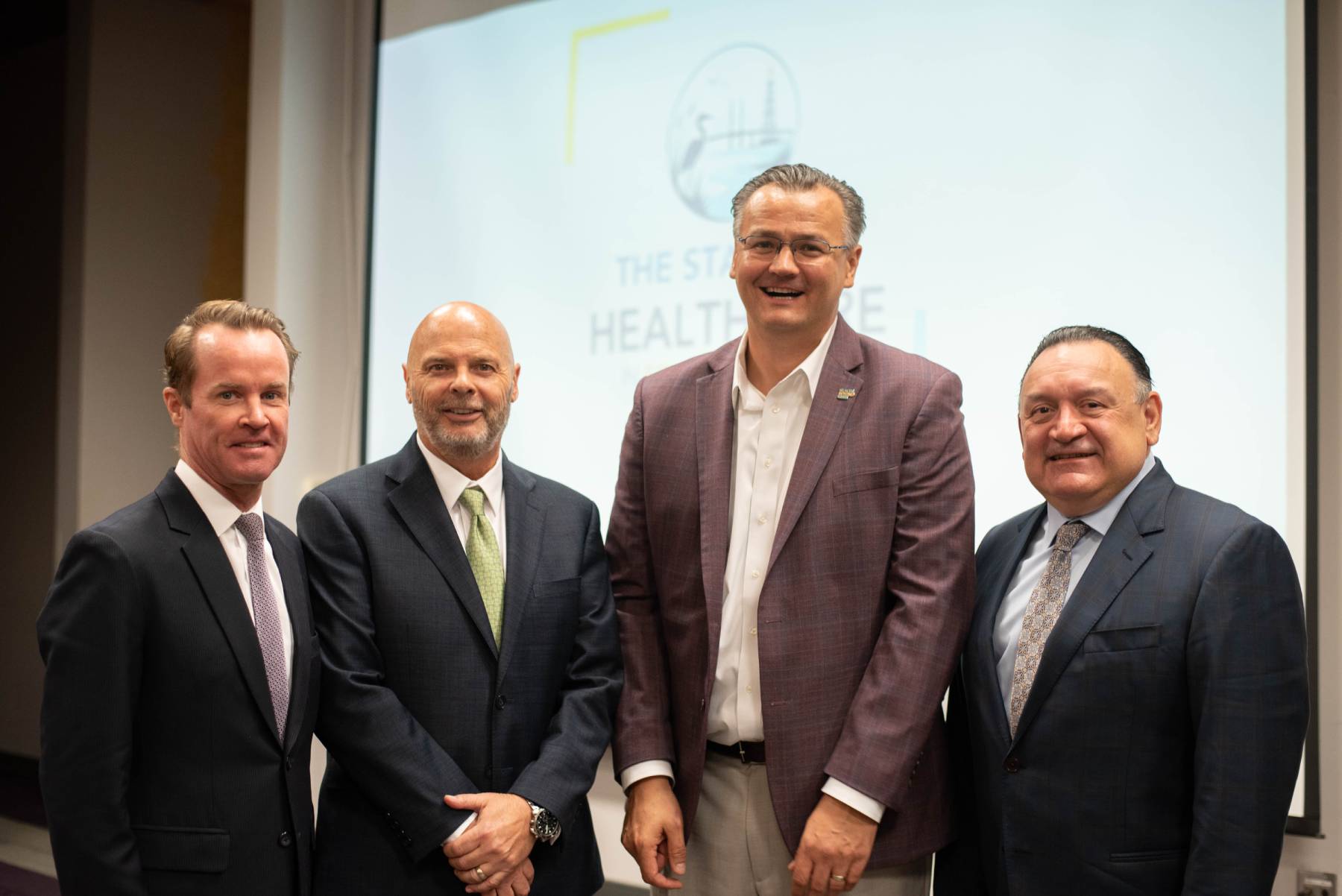 Four people stand together in front of a projection screen displaying the State of Healthcare event logo.