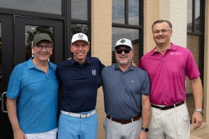Four people stand closely together smiling, dressed in golf attire. The person on the right wears a shirt with "Legacy" embroidered on it. They are in front of a building with large windows.